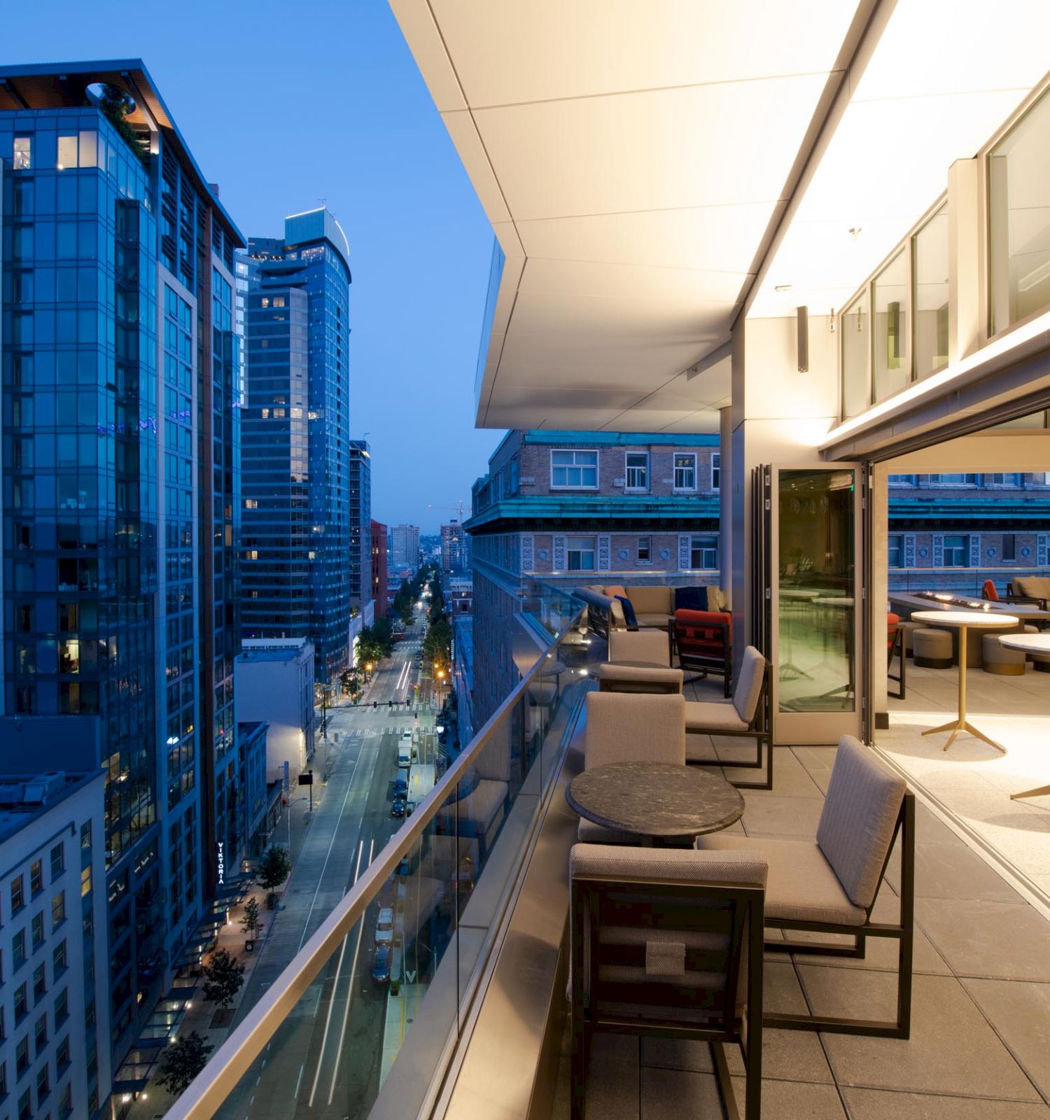 A modern balcony with tables and chairs overlooks a city skyline with tall buildings at dusk.