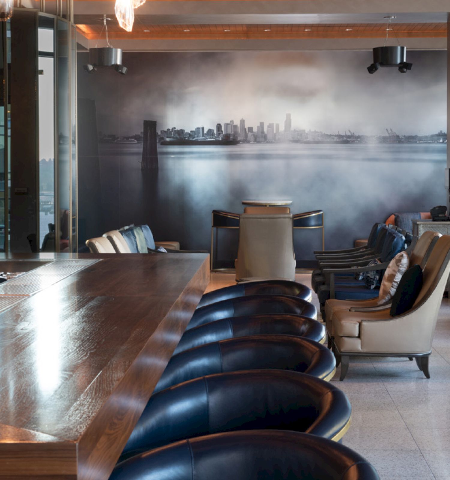 The image shows a modern bar with a wooden counter, blue chairs, and lounge seating. There is a large cityscape mural on the wall in the background.