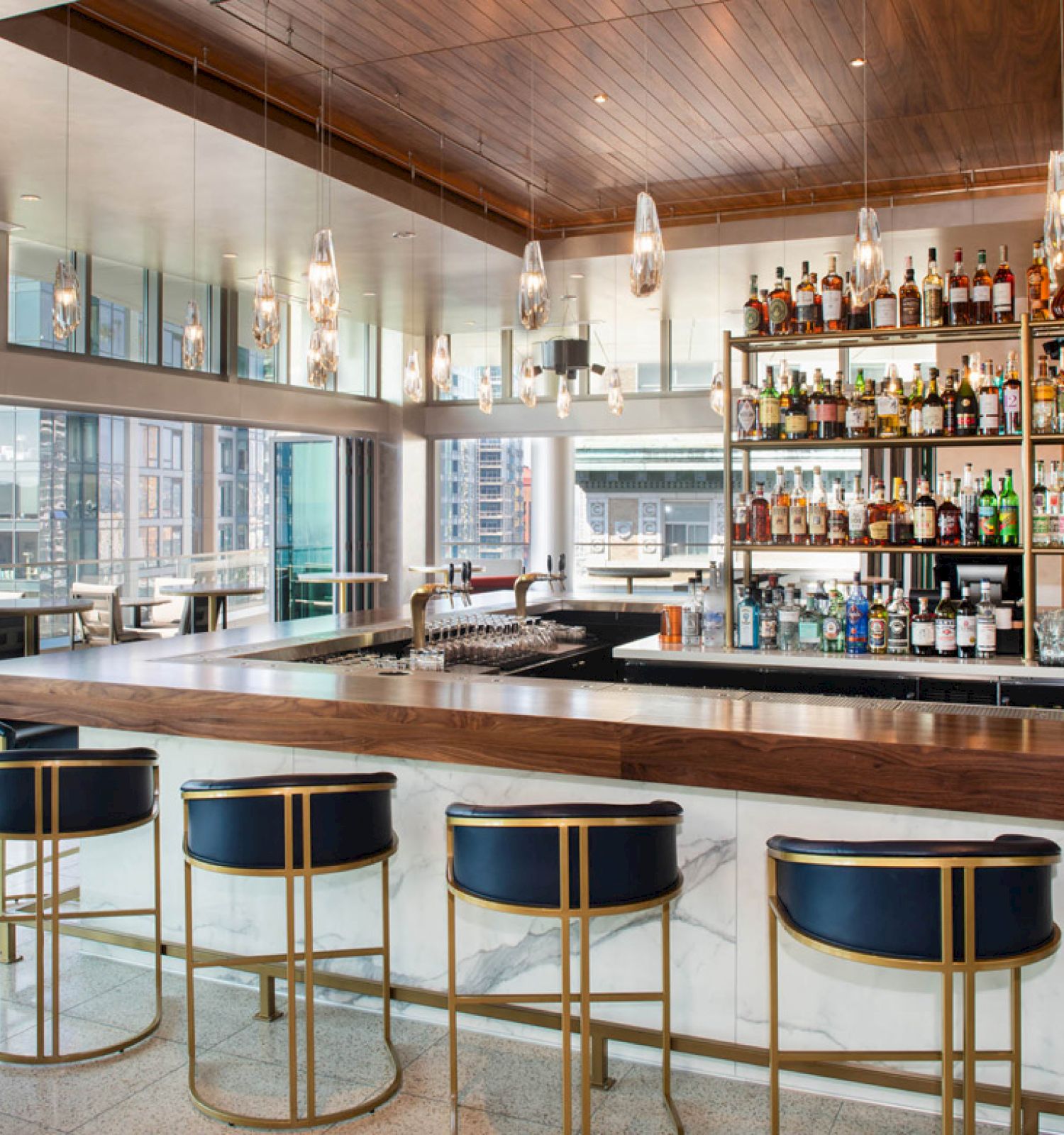 The image shows a modern bar with high stools, a well-stocked bar shelf, large windows, and a chic aesthetic in a high-rise setting.