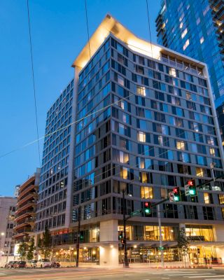 The image shows a modern high-rise building at dusk, with illuminated windows and streetlights, located at a city intersection.