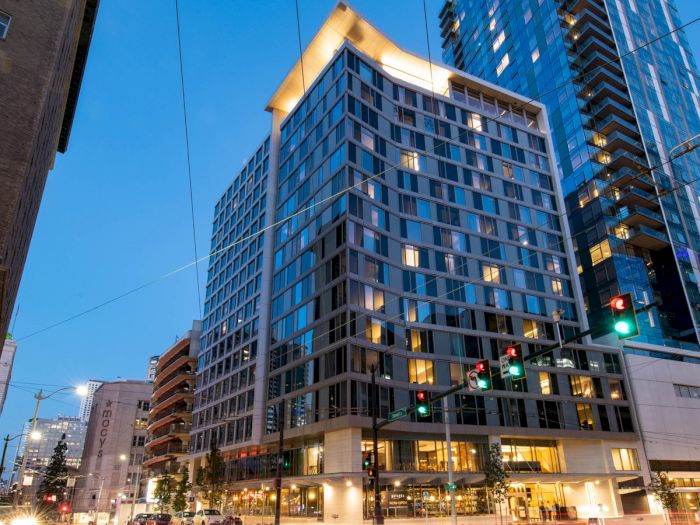 The image shows a modern high-rise building at dusk, with illuminated windows and streetlights, located at a city intersection.