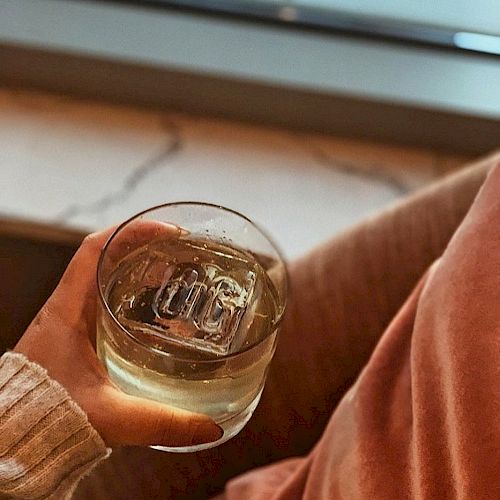 A person is holding a glass of a clear beverage with ice, while seated in a cozy environment, possibly on a couch, with a window in the background.