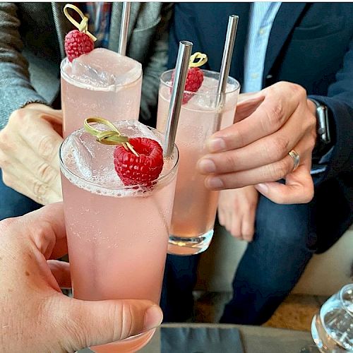 Three people are toasting with pink beverages in tall glasses, garnished with raspberries and ice cubes.