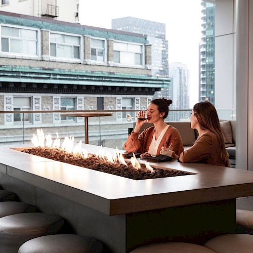 Two people are sitting at a fire table on a rooftop patio with city buildings in the background.