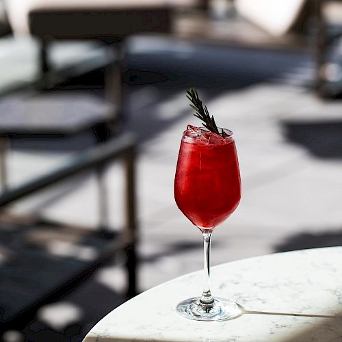 A red cocktail garnished with a sprig of rosemary is on a marble surface, with blurred outdoor seating in the background.