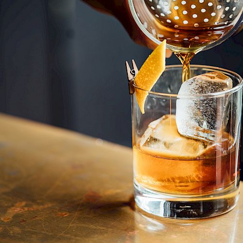 A drink is being poured into a glass with ice and a citrus peel garnish on a bar counter.