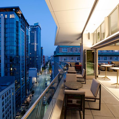 A modern urban balcony at twilight overlooks city buildings, featuring outdoor seating, tables, and open glass doors leading to the interior.