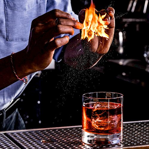 A person is preparing a cocktail, igniting a citrus peel over the drink, which is in a glass on a bar counter.