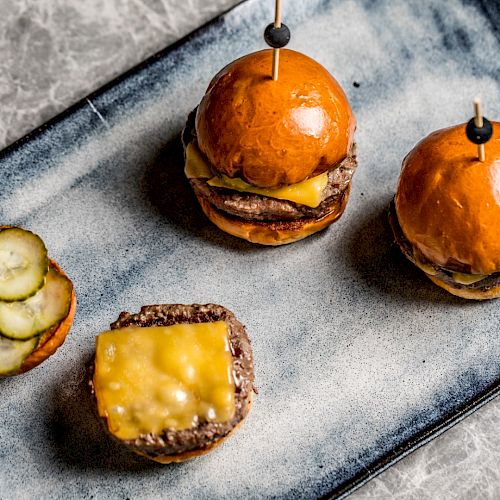 The image shows three mini cheeseburgers on a rectangular plate: two assembled and one open, revealing cheese and pickles on the buns.