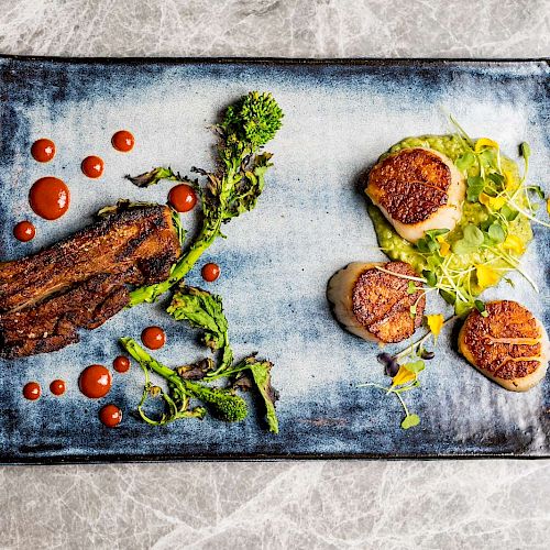 A gourmet dish on a rectangular plate, featuring seared scallops, greenery, and a piece of meat with red sauce dots.