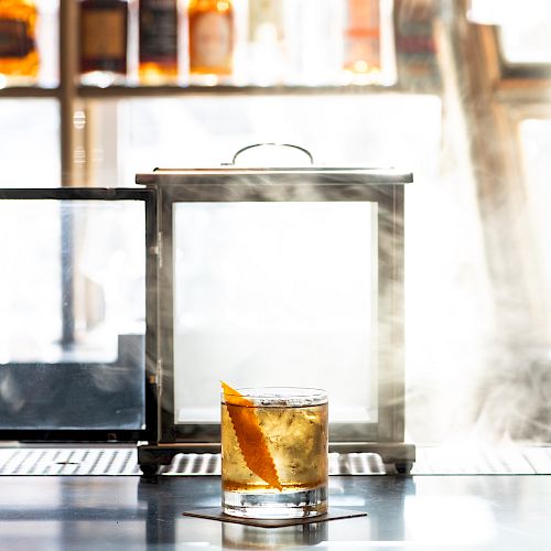 A smoky bar setup featuring a glass of whiskey garnished with an orange peel, placed in front of a glass box emitting smoke on a bar counter.