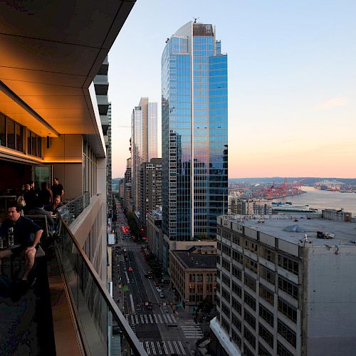 People are on a balcony overlooking a city street with tall buildings and a scenic view of a river in the background at sunset.