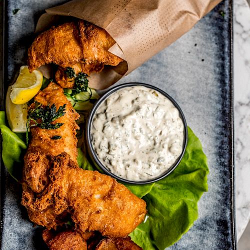 The image shows a plate with golden-brown fried fish, a small bowl of tartar sauce, garnished with lettuce and a lemon wedge, and brown parchment paper.