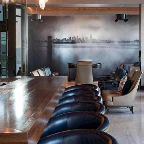 A modern, stylish bar with a long wooden counter, blue bar stools, and cozy seating arrangements. A cityscape mural decorates the back wall.