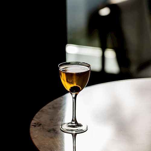 A glass of amber liquid, possibly whiskey or wine, sits on a reflective circular table in a dimly lit setting.