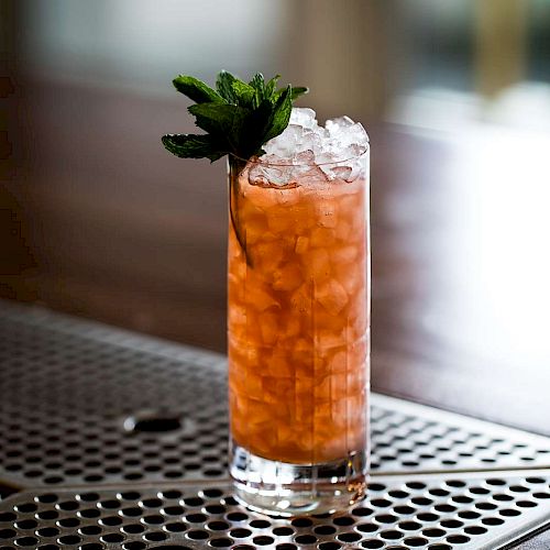 The image shows a tall glass filled with a reddish-orange iced drink, topped with mint leaves, placed on a perforated metal surface.