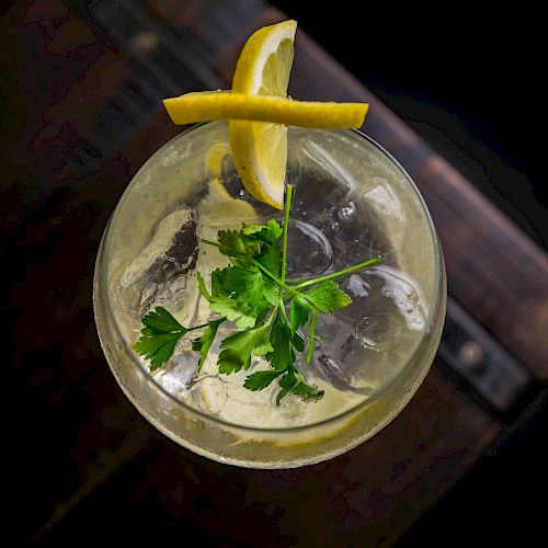 A cocktail garnished with lemon slices and a sprig of parsley in a round glass, viewed from above, on a dark wooden surface.