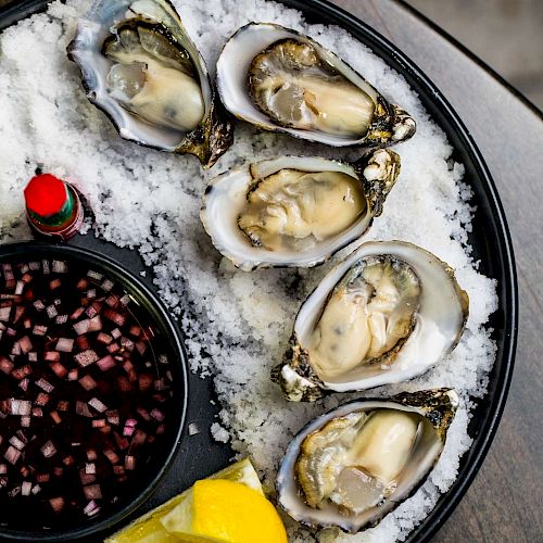 A plate of oysters on a bed of ice, with lemon wedges, a small bottle of hot sauce, and a bowl of diced onions and vinegar.