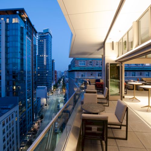 The image shows a modern urban balcony with tables and chairs, overlooking a cityscape with tall buildings at dusk.