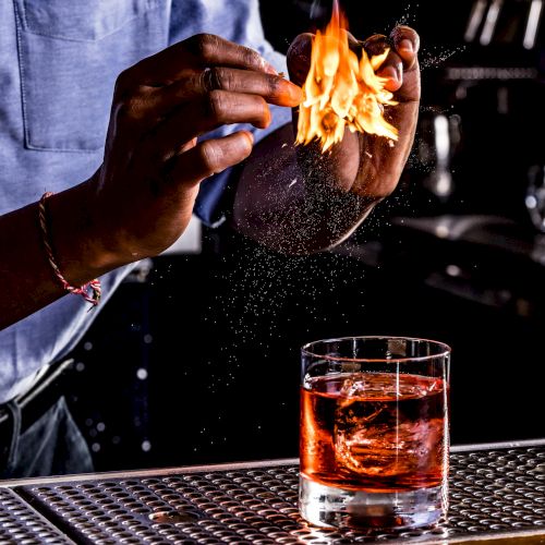 A person is igniting an orange peel over a cocktail glass, creating a burst of flames above the drink, likely for added aroma and visual effect.