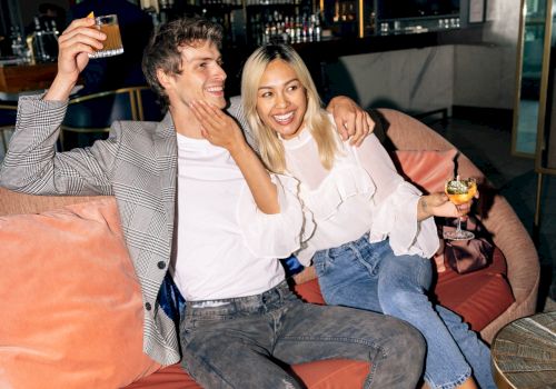 A smiling couple relax on a lounge sofa in a bar, each holding a cocktail as warm lights glow behind them.