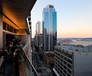 City skyline seen from a balcony at sunset, with office workers nearby and a tall glass skyscraper by a waterfront harbor.