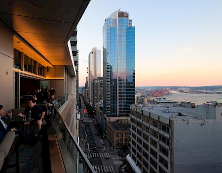 City skyline seen from a balcony at sunset, with office workers nearby and a tall glass skyscraper by a waterfront harbor.