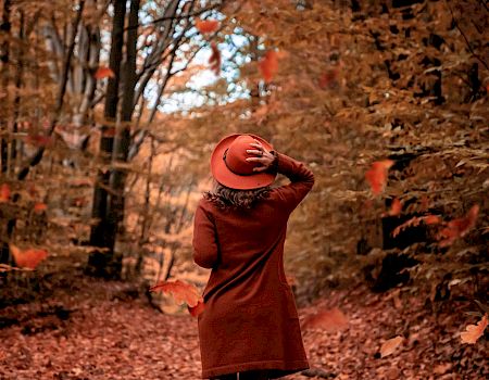 A person in an autumn forest in a red hat throws orange leaves into the air, smiling through the falling fall foliage.