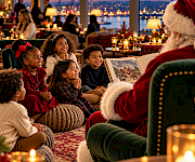 Santa reading a holiday storybook to smiling children on floor cushions with a candlelit Seattle waterfront view at sunset