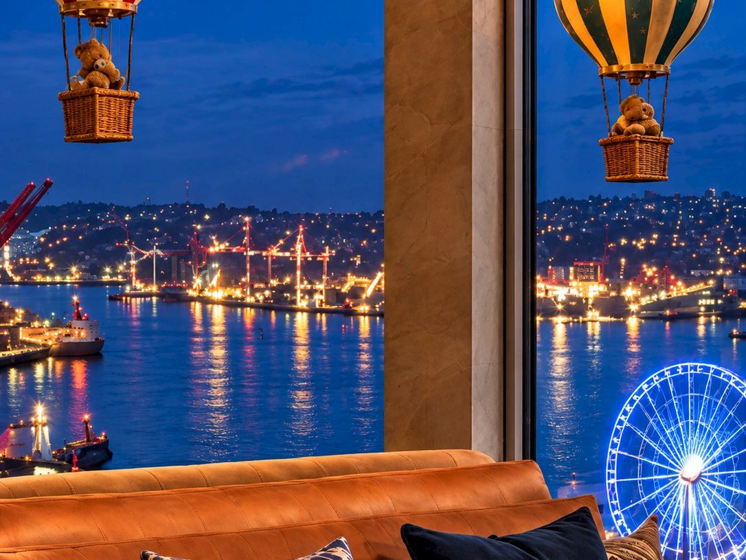 A cozy living room overlooks a night harbor, with glowing hanging bulbs, balloon-themed lanterns, and a blue-lit Ferris wheel.