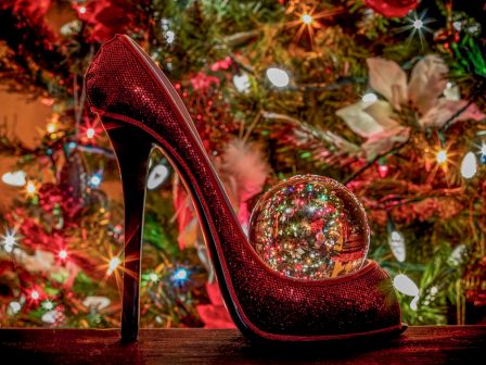 A shiny red high-heeled shoe with a reflective ornament sits by a glittering Christmas tree, with colorful lights and star-like bokeh.